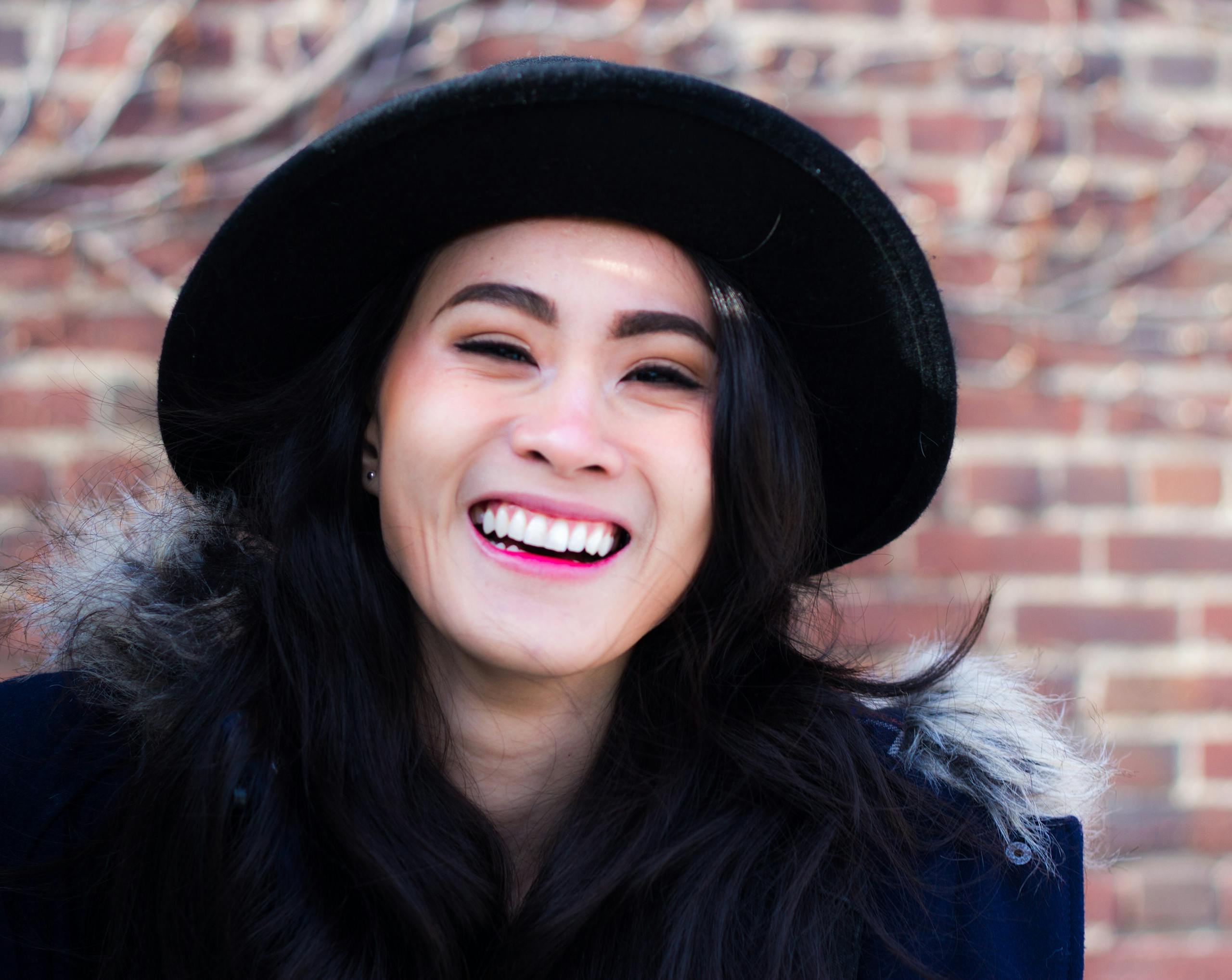 Happy woman wearing a black hat and fur-trimmed coat, laughing outdoors against a brick wall.